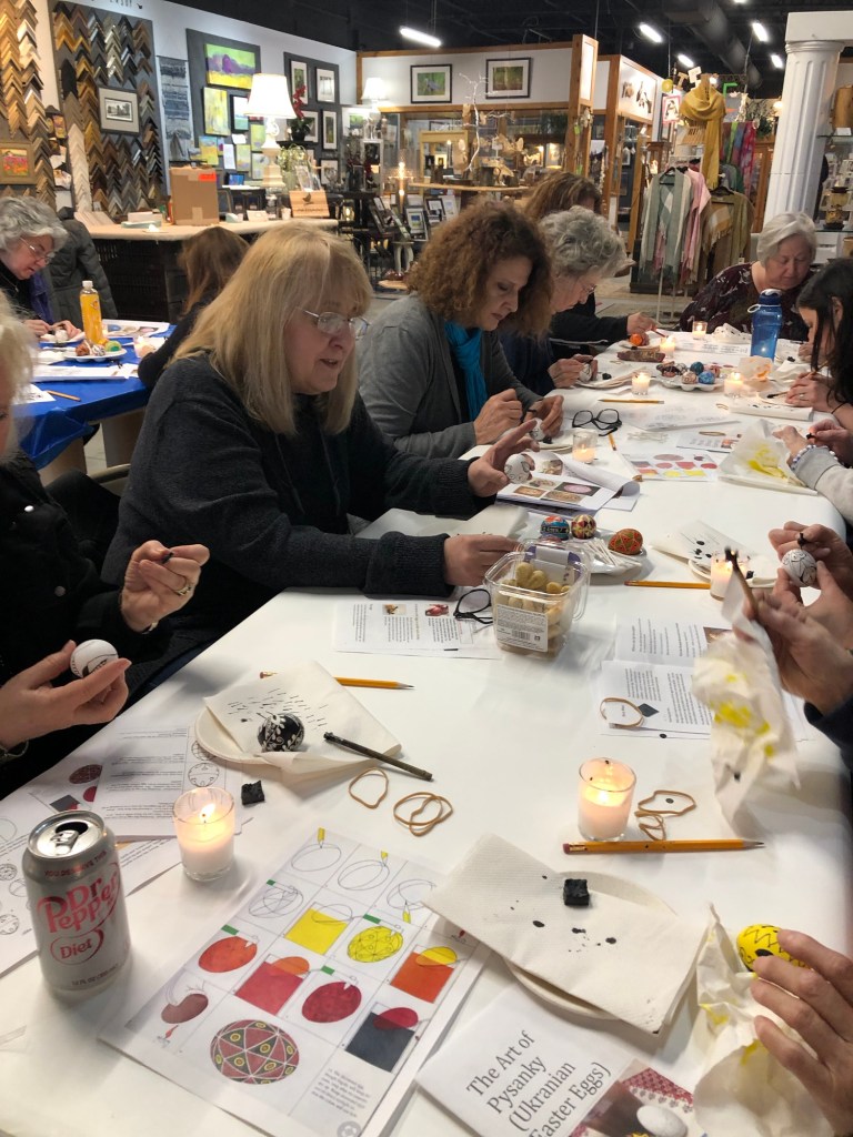 A group of women engaged in a painting workshop, creating decorative eggs. The participants are seated at a table surrounded by art supplies, candles, and instructional materials.