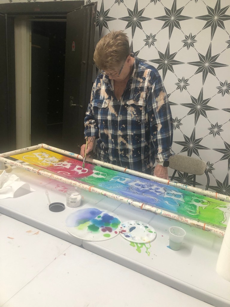 An artist is painting colorful designs on a long canvas while surrounded by various paint supplies. The background features a patterned wall.