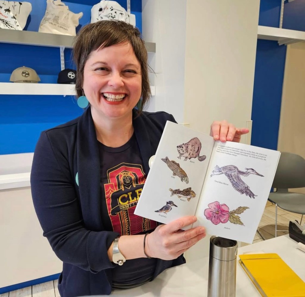 A smiling woman holds an open book showcasing illustrations of animals and plants, while standing in a brightly colored room with merchandise displayed in the background.