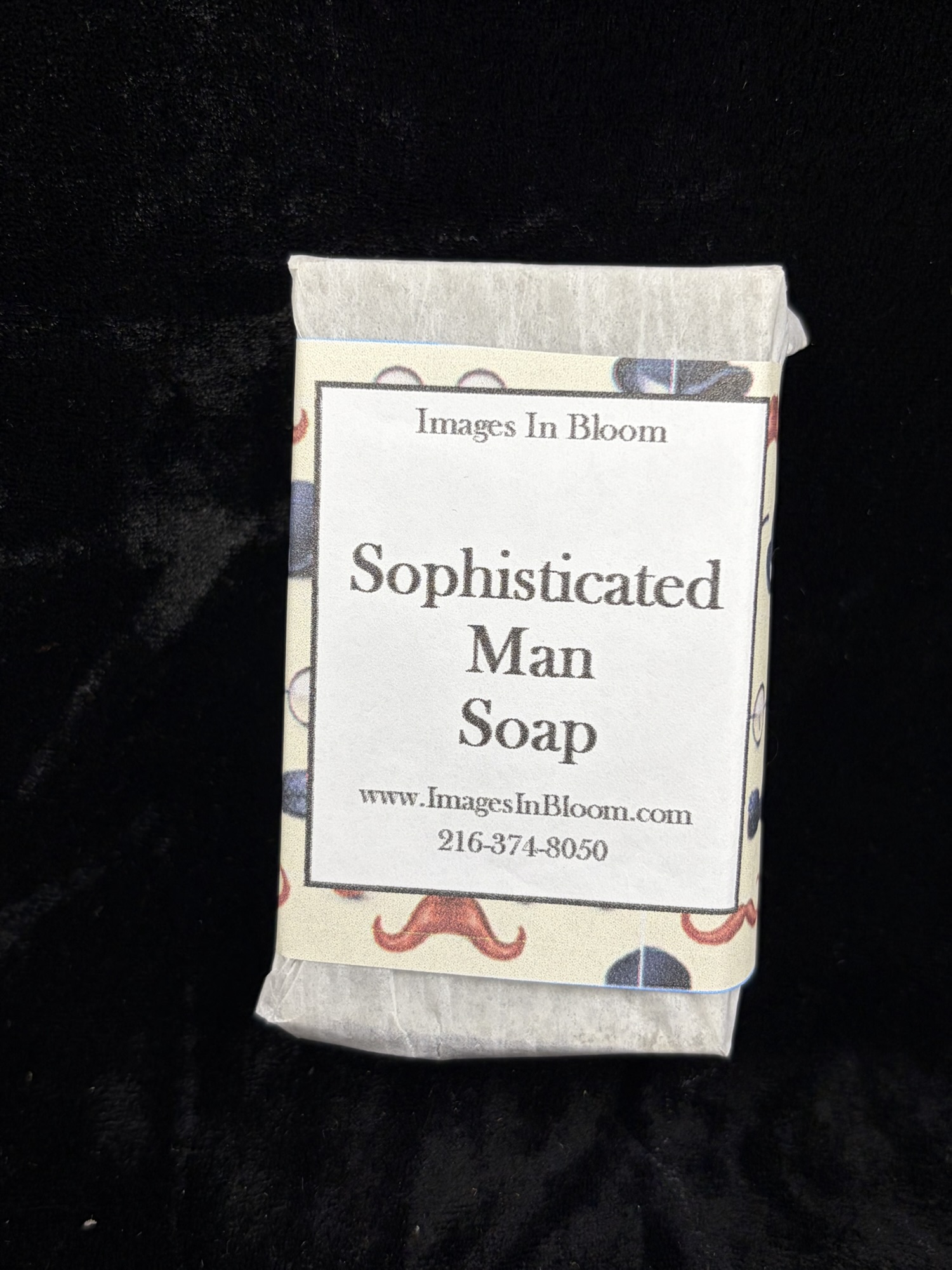 Bar of soap labeled 'Sophisticated Man Soap' from Images In Bloom, featuring decorative packaging with patterns and contact information.