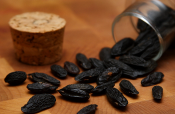 A glass jar tipped over, spilling black seeds onto a wooden surface, with a cork lid nearby.
