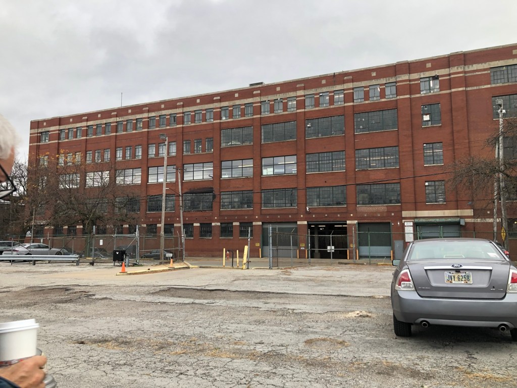 A large red brick industrial building with many windows, viewed from a parking lot. A grey car is parked in the foreground, and there is fencing and construction equipment around the entrance.