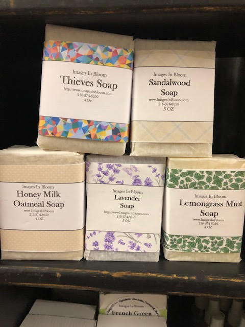 Five bars of soap displayed on a shelf, labeled as Thieves Soap, Sandalwood Soap, Honey Milk Oatmeal Soap, Lavender Soap, and Lemongrass Mint Soap, each with distinct packaging.