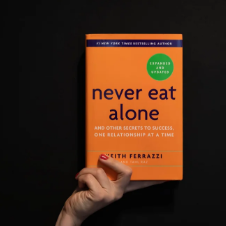 A hand holding the book 'Never Eat Alone' by Keith Ferrazzi, featuring an orange cover with the subtitle 'And Other Secrets to Success, One Relationship at a Time'.