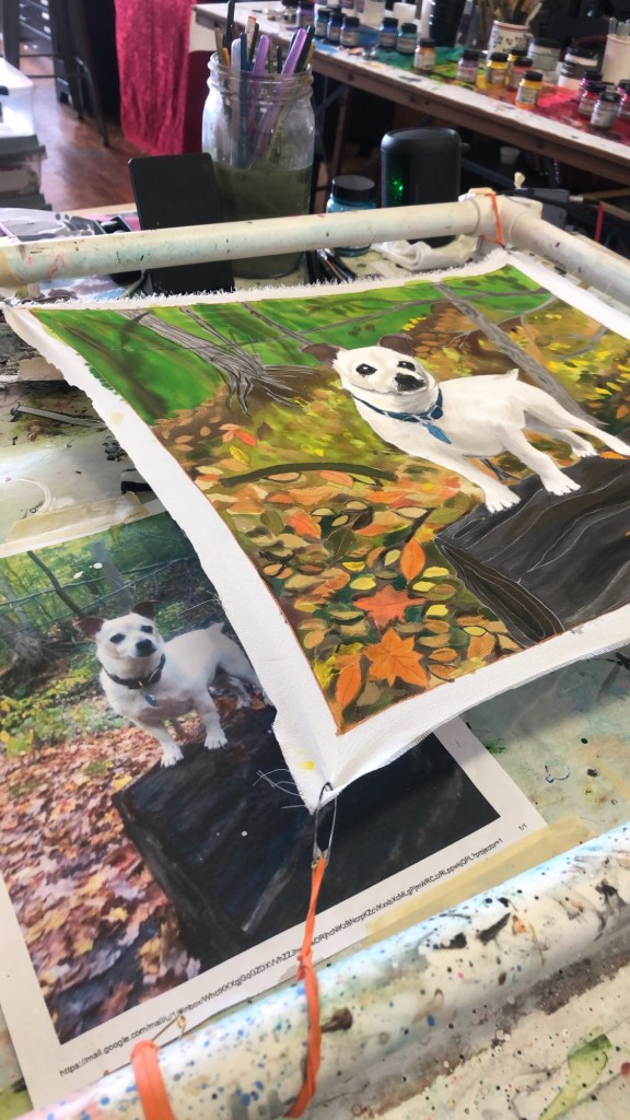 A painted canvas featuring a dog standing on a log surrounded by autumn leaves, alongside a printed photo reference of the same scene.