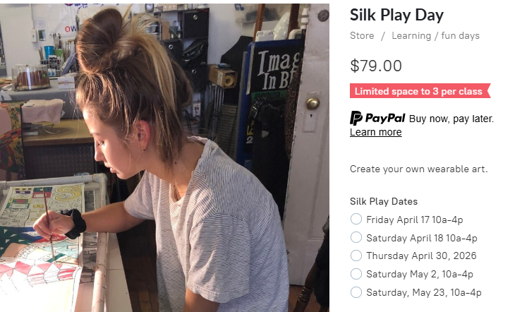A young woman with a bun hairstyle is focused on painting a colorful design on a silk material in a creative studio setting, with various art supplies visible in the background.
