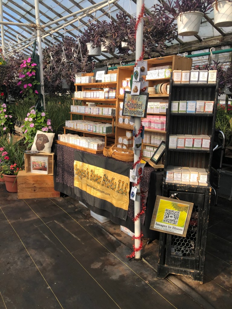 A vibrant greenhouse display featuring a craft booth. The booth has shelves filled with various products, including wire crochet brooches. Bright hanging plants and flowers adorn the background, creating a lively atmosphere.