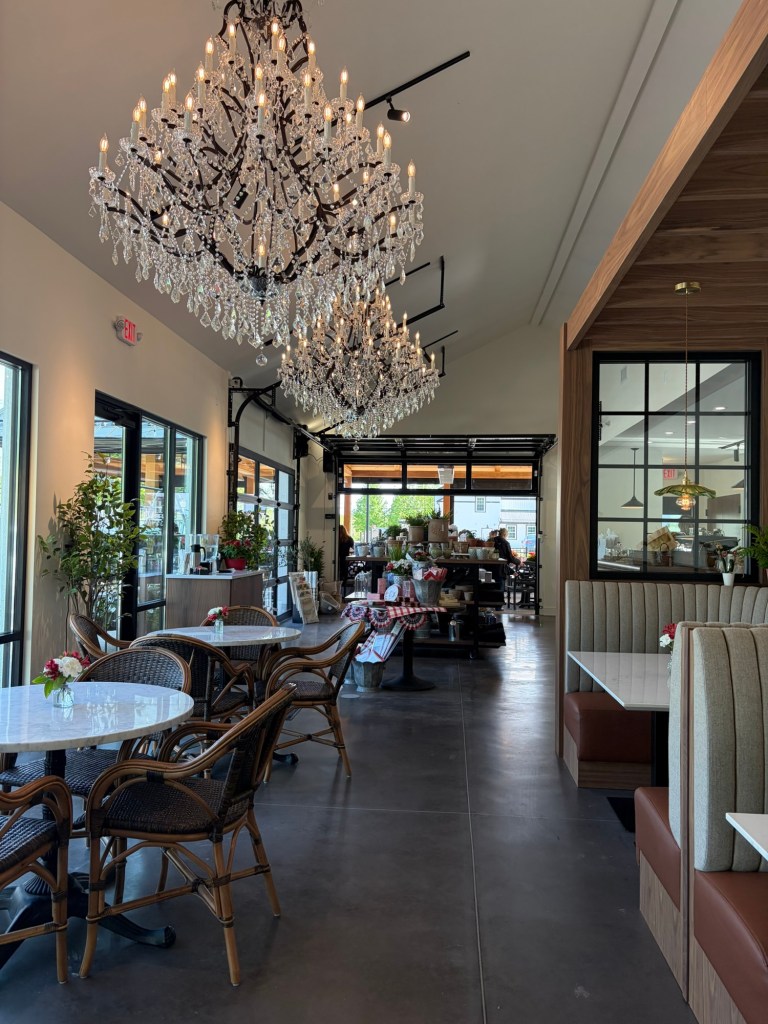 Interior of a stylish cafe featuring elegant chandeliers, wooden furnishings, and cozy seating arrangements with small flower decorations on tables.