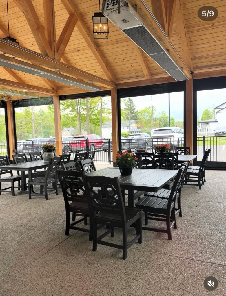 Outdoor dining area with wooden beams, multiple black tables and chairs, and potted plants.